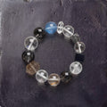 Z01 Ferro Barrier bracelet — black super seven, rutilated and clear quartz beads with one aquamarine, flat lay on dark stone